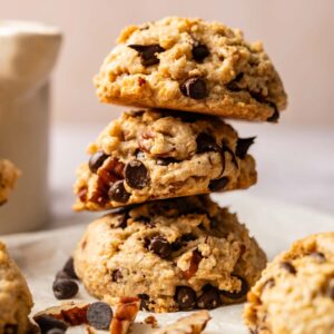 Three Cowboy Cookies stacked on parchment paper, with chocolate chips and pecan pieces scattered nearby; a white cup is blurred in the background.