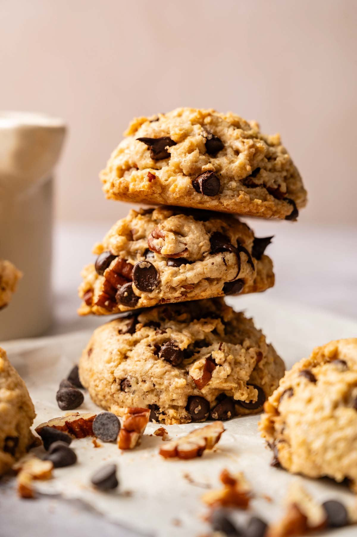 Three Cowboy Cookies stacked on parchment paper, with chocolate chips and pecan pieces scattered nearby; a white cup is blurred in the background.