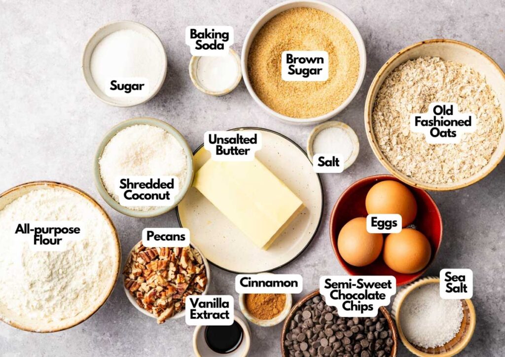 Various labeled baking ingredients in bowls, including all-purpose flour, sugars, oats, butter, eggs, coconut, pecans, vanilla, cinnamon, chocolate chips, salt, and baking soda—perfect for making classic Cowboy Cookies.