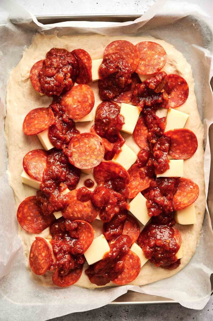 Detroit Style Pizza features rectangular dough topped with cubed cheese, sliced pepperoni, and tomato sauce on a parchment-lined baking tray.