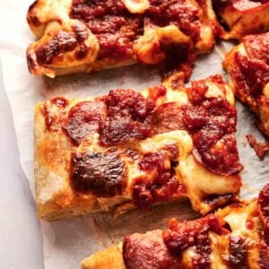 Rectangular slices of Detroit Style Pizza feature thick crust, melted cheese, and chunky tomato sauce, all served on parchment paper.