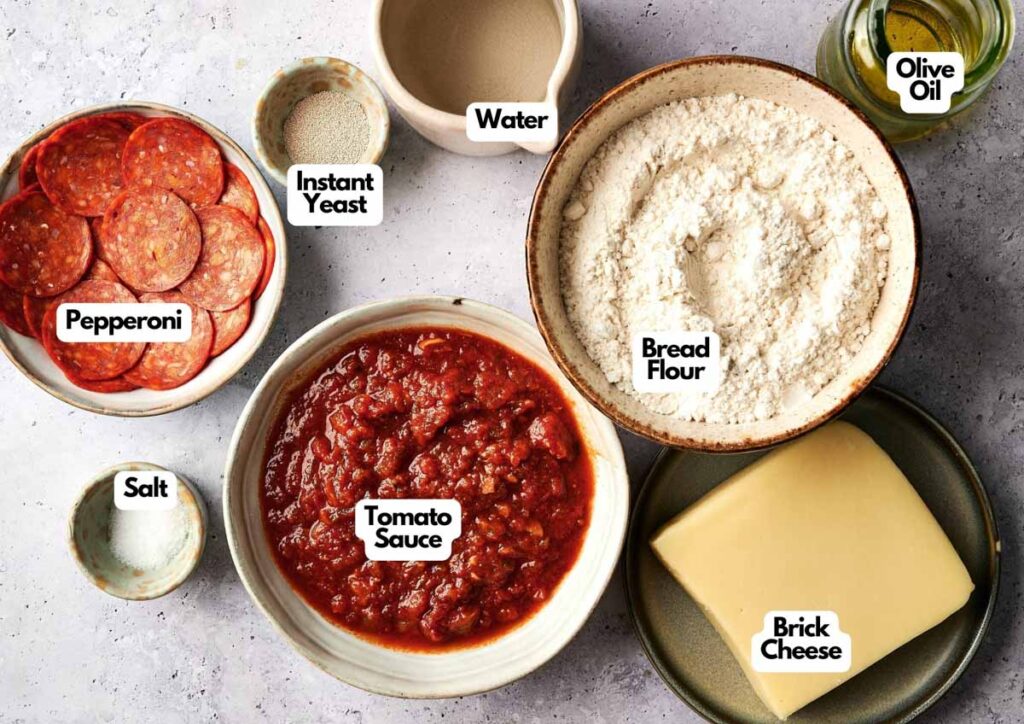 Various Detroit Style Pizza ingredients labeled on a counter, including pepperoni, instant yeast, water, bread flour, olive oil, salt, tomato sauce, and brick cheese.