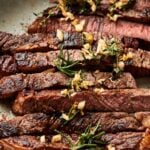 Sliced reverse sear steak topped with chopped garlic and fresh herbs on a plate.