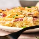 A close-up of a slice of scalloped potatoes and ham gratin with cheese, garnished with chopped parsley, served on a white plate with a fork.