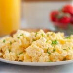 A plate of Scrambled Eggs with Cream Cheese garnished with chopped chives, with out-of-focus strawberries and a glass of orange juice in the background.