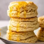 A stack of three golden sourdough biscuits with honey dripping down the sides, placed on a plate with more biscuits in the background.