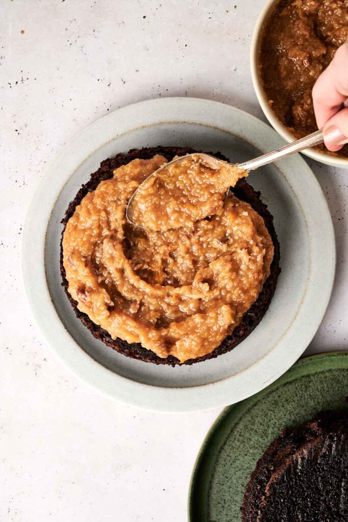 A hand spreads a thick brown mixture on a dark cake layer with a spoon, on a light-colored plate. A bowl with more mixture is in the background.