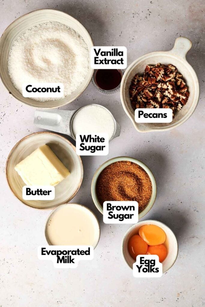 Various baking ingredients are arranged on a surface: coconut, pecans, butter, evaporated milk, vanilla extract, white sugar, brown sugar, and egg yolks.