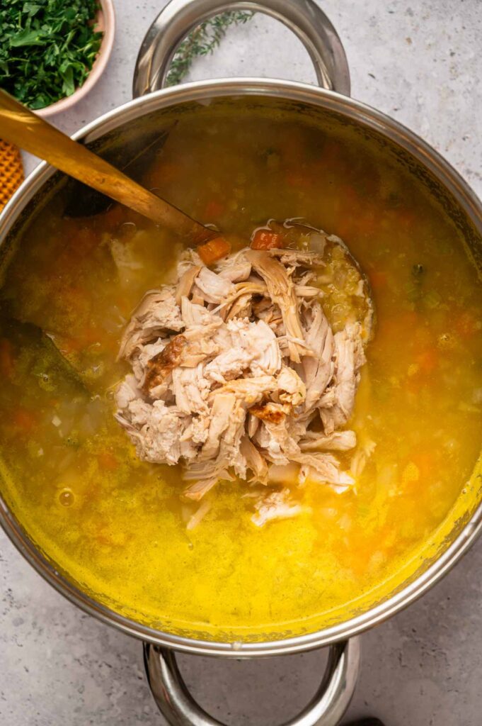 A pot of Italian Penicillin Soup with shredded chicken being added, surrounded by diced vegetables in a rich yellow broth.