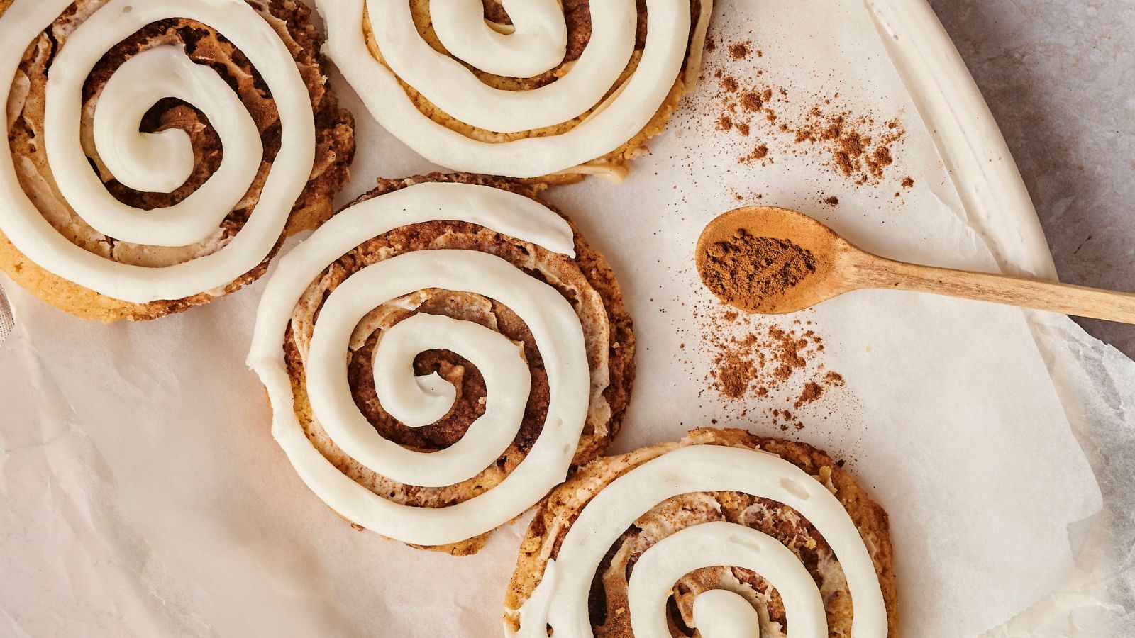 Four cinnamon rolls with white icing sit on parchment paper beside a wooden spoon containing ground cinnamon.