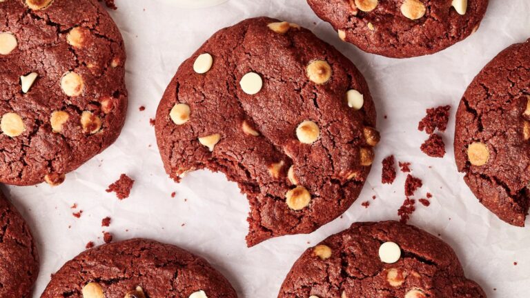 Several red velvet cookies with white chocolate chips are shown on parchment paper, with one cookie in the center missing a bite.