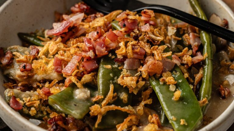 A bowl of green beans and snap peas topped with chopped bacon, crispy fried onions, and a creamy sauce, with a black spoon resting on the edge.