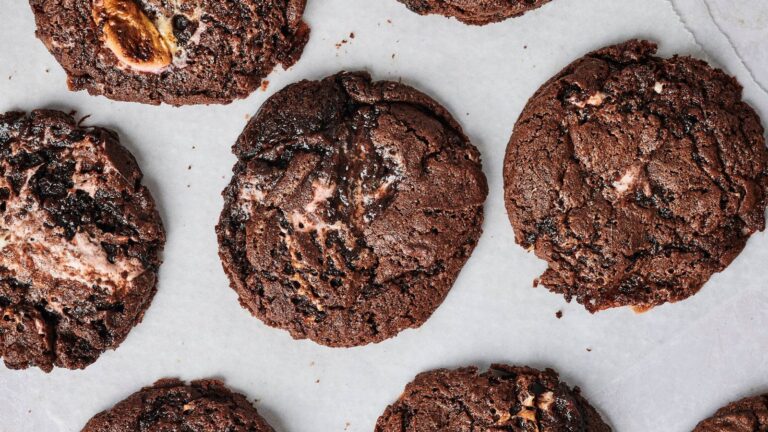 Several chocolate cookies with visible chunks and melted marshmallow pieces are arranged on a sheet of parchment paper.