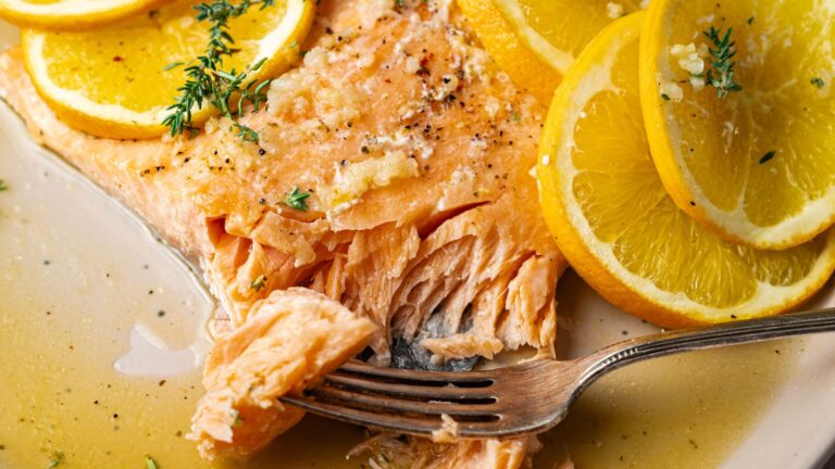 A fork flakes a cooked salmon fillet topped with lemon slices and herbs, with sauce pooling on the plate.