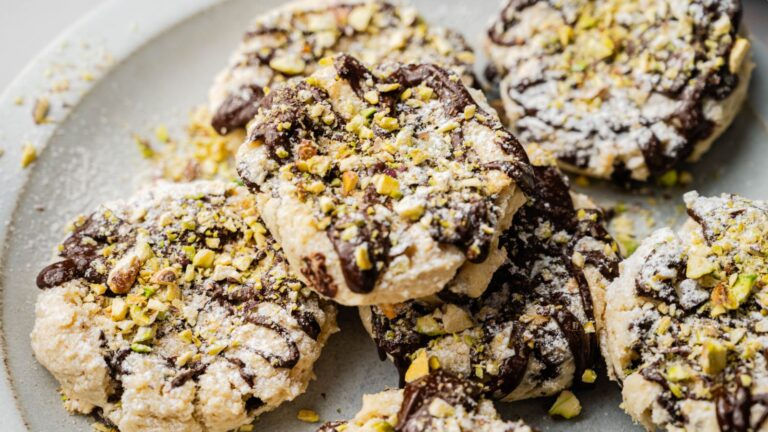 A plate of cookies topped with powdered sugar, chopped pistachios, and drizzled with chocolate.