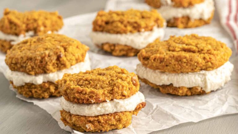 Five pumpkin whoopie pies with cream filling are arranged on pieces of parchment paper on a light-colored surface.