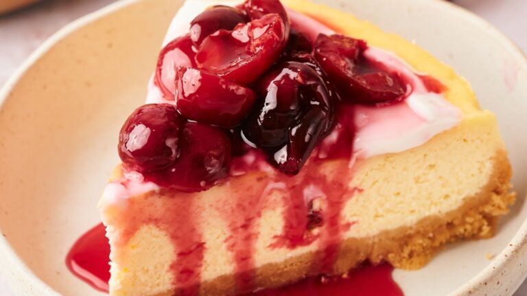 A slice of cheesecake topped with cherry compote sits on a beige plate, with red sauce dripping down the sides.