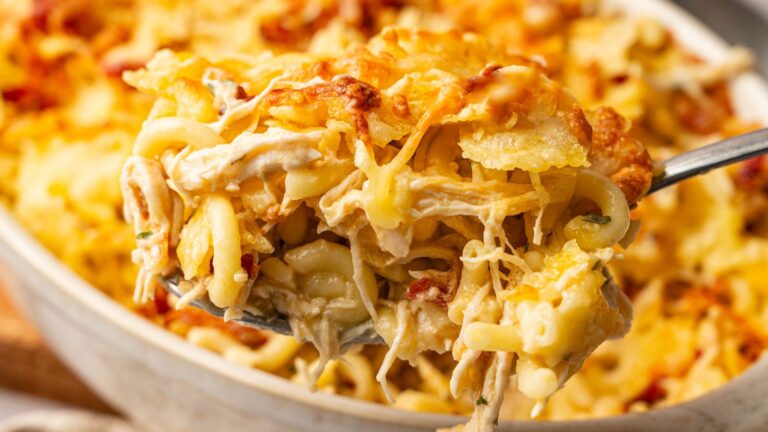 Close-up of a spoonful of cheesy baked pasta casserole with shredded chicken and melted cheese, held above a dish.