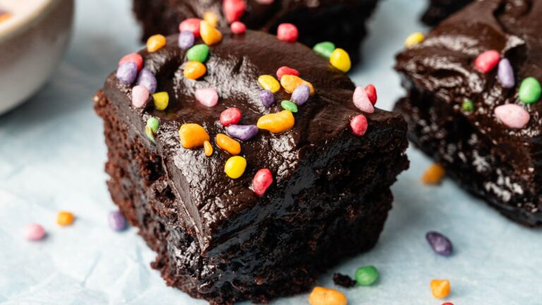 A close-up of a chocolate brownie topped with colorful candy sprinkles, placed on a sheet of parchment paper.