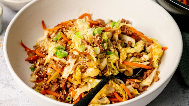A bowl of stir-fried ground meat, shredded cabbage, carrots, and green onions, topped with sesame seeds, with a black spoon in the dish.