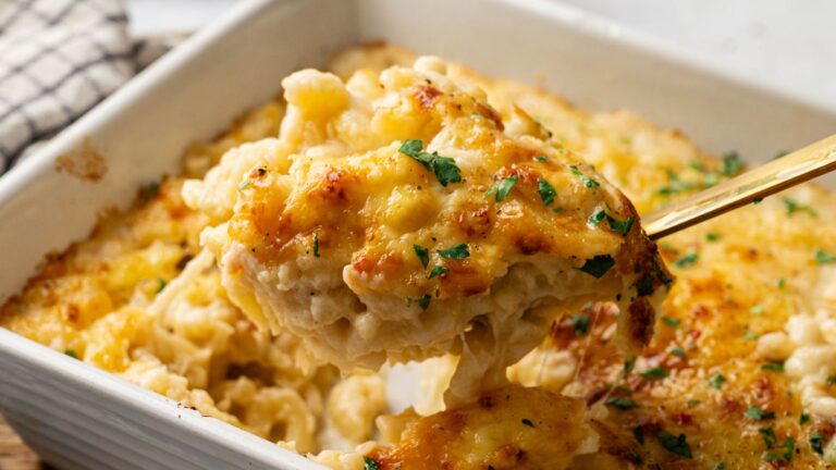 A serving spoon lifts a portion of baked macaroni and cheese topped with melted cheese and garnished with chopped parsley from a casserole dish.