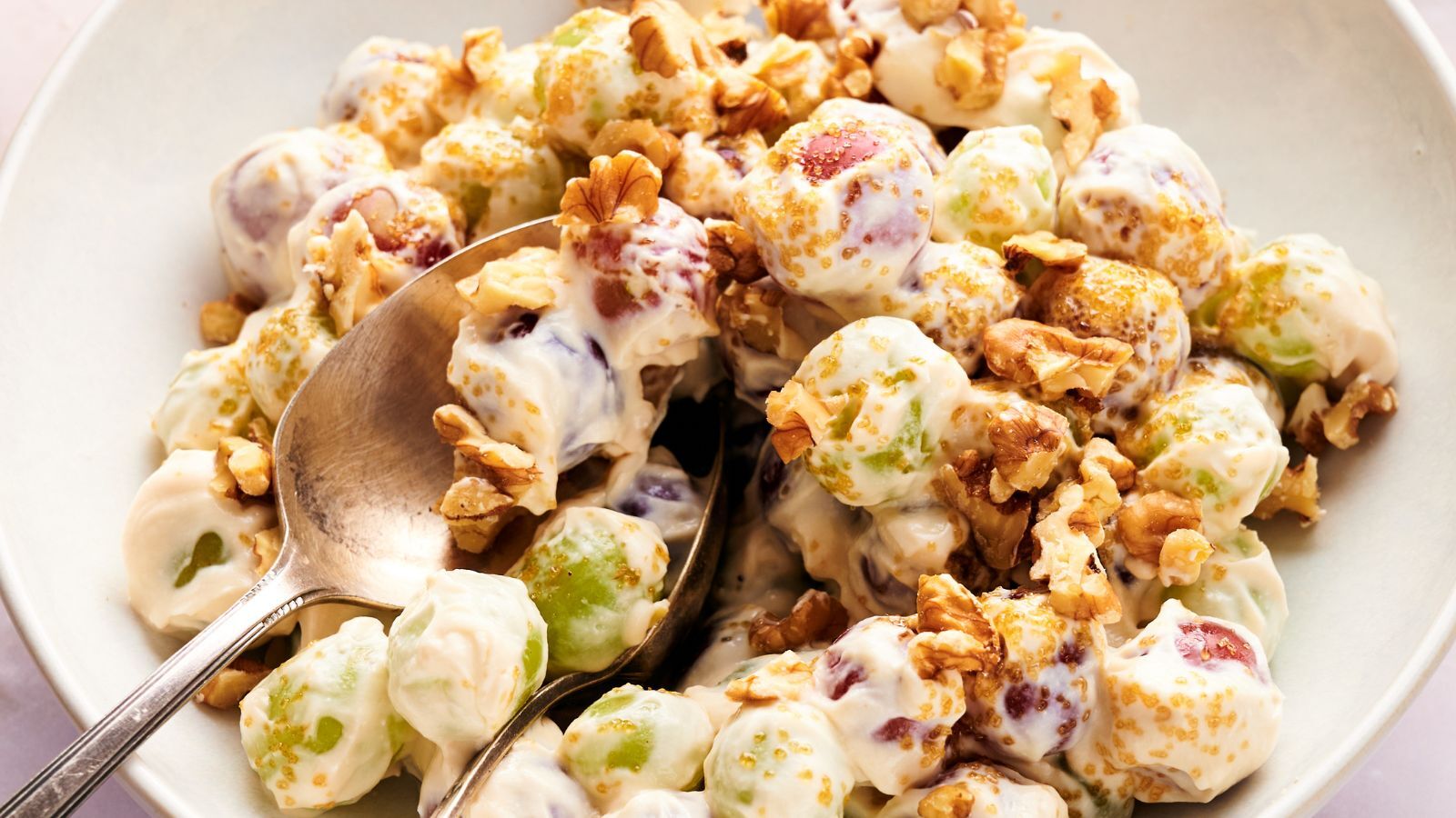 A bowl of grape salad with green and red grapes coated in creamy dressing, topped with chopped walnuts and brown sugar, with a spoon for serving.