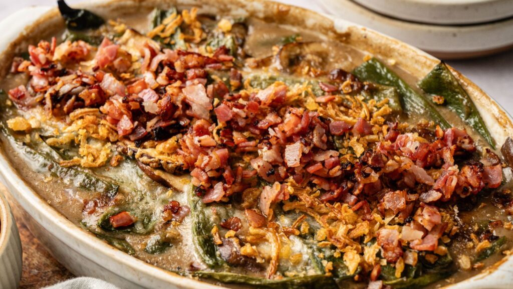 A baked casserole topped with chopped bacon, crispy onions, and green vegetables in a creamy sauce, served in a white oval dish.