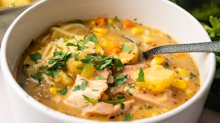 A bowl of creamy chicken and vegetable soup with potatoes, corn, carrots, and fresh herbs, served with a spoon.