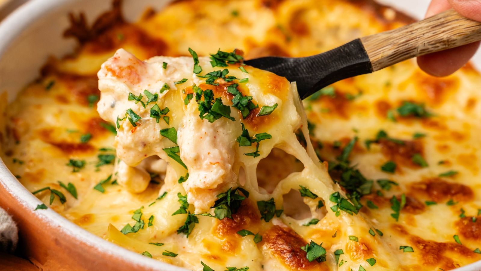 A spoon lifts a portion of cheesy baked pasta topped with chopped parsley from a casserole dish.