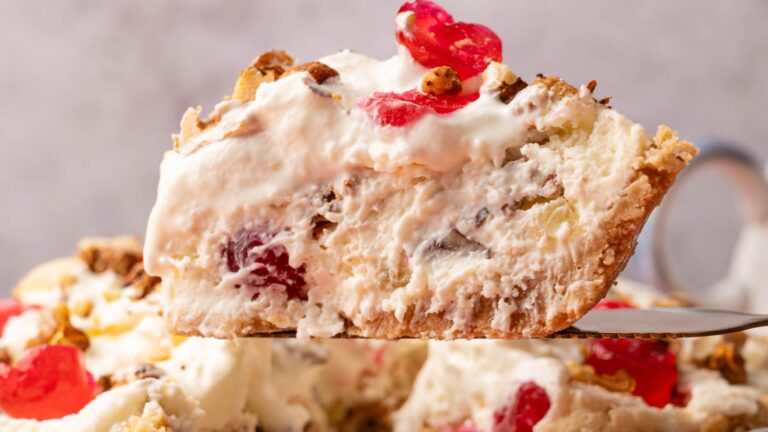 A close-up of a slice of creamy pie with whipped topping, chopped nuts, and maraschino cherries on a pie server.