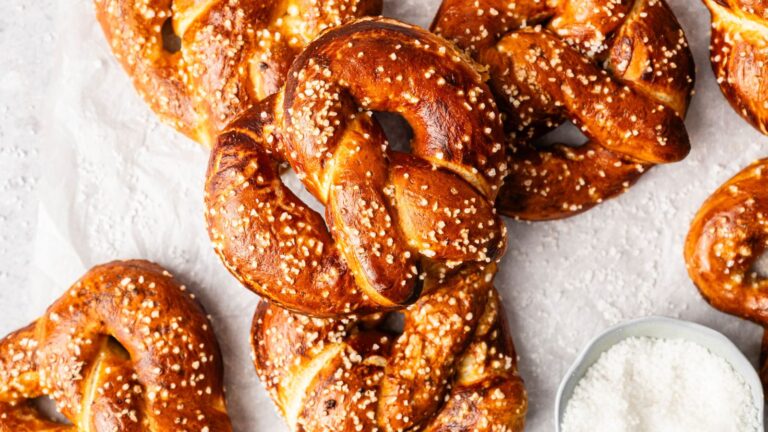 Several golden-brown soft pretzels sprinkled with coarse salt are arranged on a light surface next to a small bowl of salt.