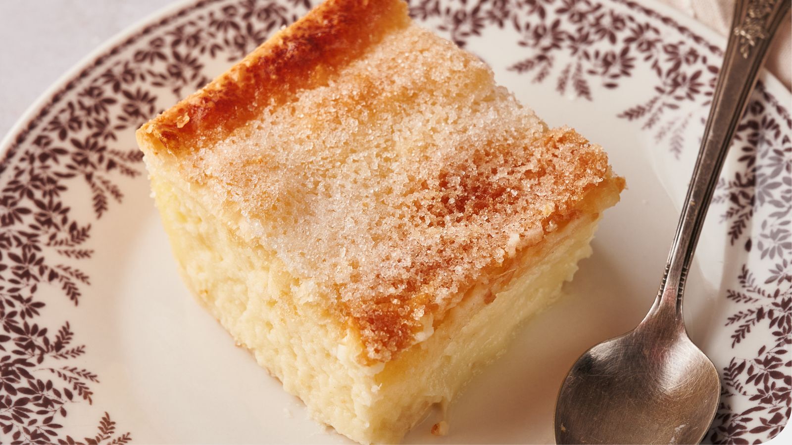 A square slice of sugar-topped cake sits on a patterned plate beside a metal spoon.