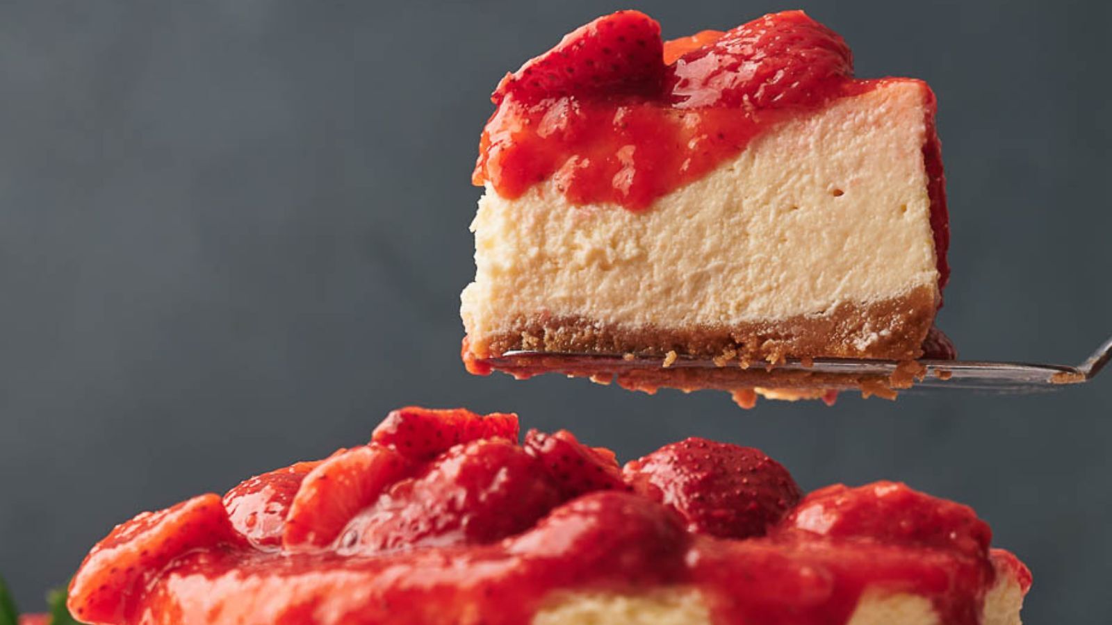 A slice of strawberry-topped cheesecake is being lifted above the rest of the cake, showing a graham cracker crust and creamy filling.