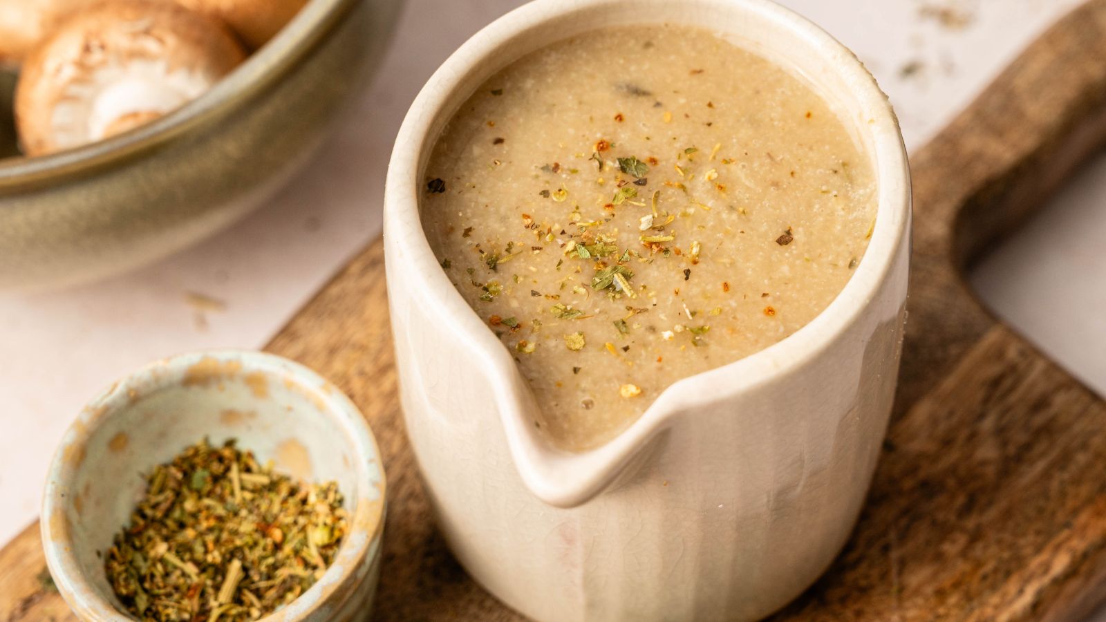 A ceramic jug filled with creamy mushroom gravy, sprinkled with herbs, sits on a wooden board next to a small bowl of mixed dried herbs.