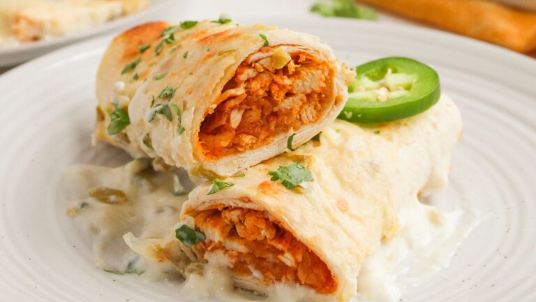 Two enchiladas filled with shredded chicken and sauce, topped with creamy white sauce, chopped cilantro, and a slice of jalapeño on a white plate.