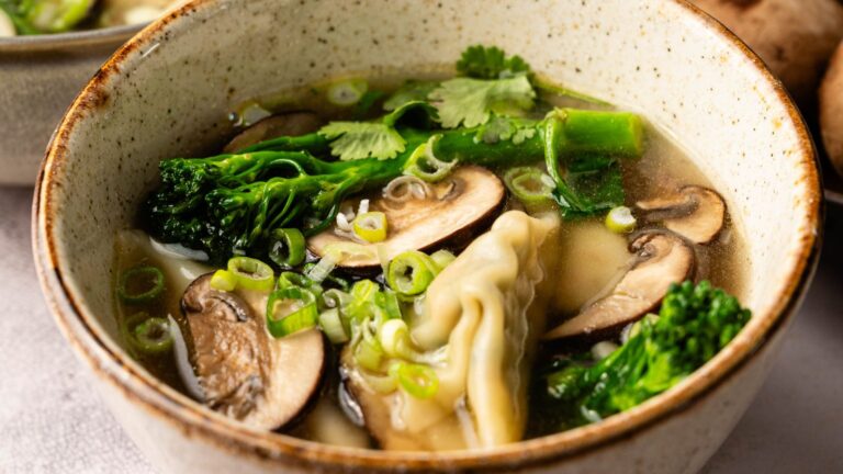 A bowl of soup with sliced mushrooms, broccoli florets, green onions, cilantro, and a dumpling in a light broth.