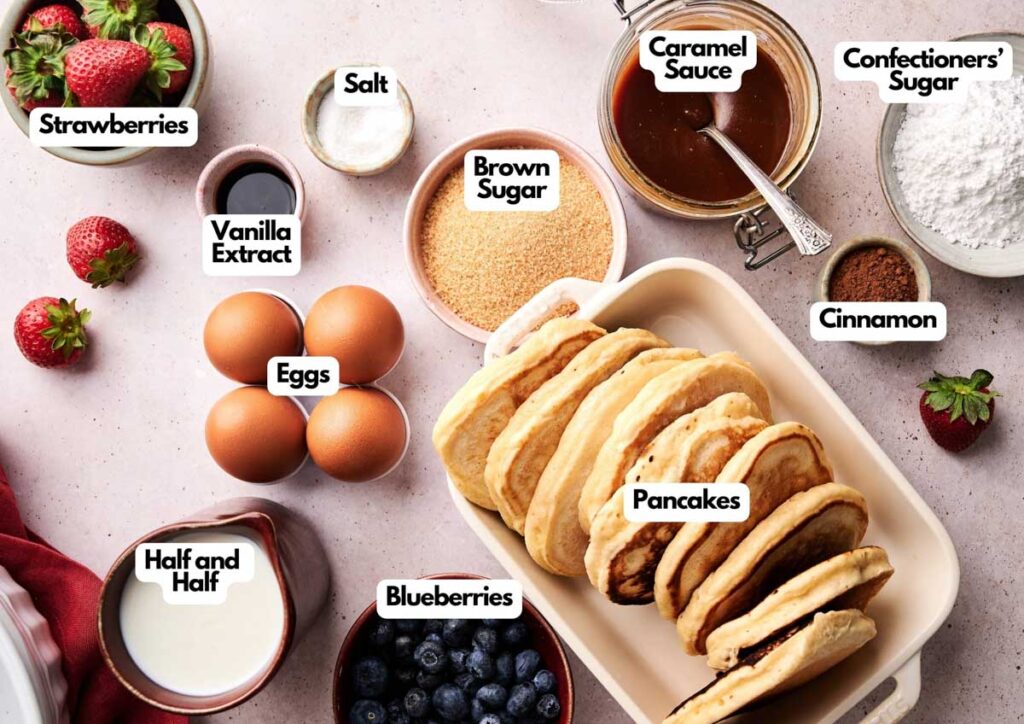 A top-down view of labeled ingredients for Air Fryer Pancake Casserole, including eggs, strawberries, blueberries, sugars, vanilla extract, cinnamon, caramel sauce, confectioners&rsquo; sugar, and half-and-half.