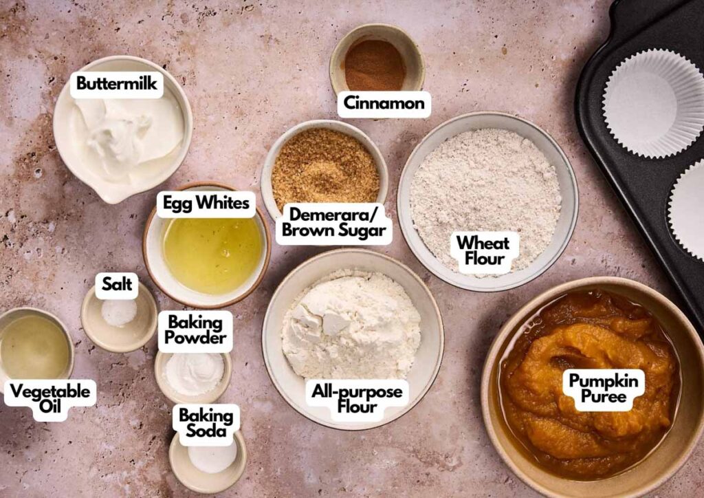 Several labeled bowls hold ingredients for baking Air Fryer Pumpkin Muffins, including buttermilk, egg whites, salt, vegetable oil, baking powder, cinnamon, brown sugar, wheat flour, all-purpose flour, and pumpkin puree.