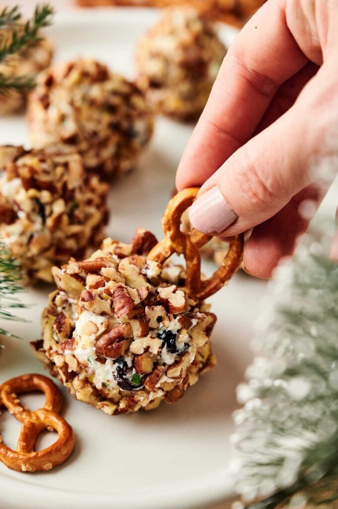 A hand holds a bite-sized Reindeer Mini Cheese Ball coated in chopped nuts, using a small pretzel as a handle. Other festive cheese balls and pretzels rest on a white plate nearby.