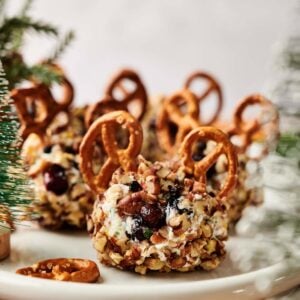 Reindeer Mini Cheese Balls coated with nuts and dried fruit, each decorated with two pretzel twists on top, are arranged on a white plate against a blurred festive background.