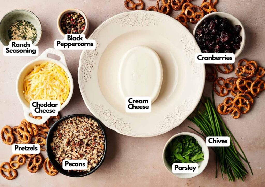 A plate with a block of cream cheese is surrounded by labeled bowls of cheddar cheese, ranch seasoning, black peppercorns, cranberries, pretzels, pecans, parsley, and chives&mdash;perfect ingredients for festive Reindeer Mini Cheese Balls.