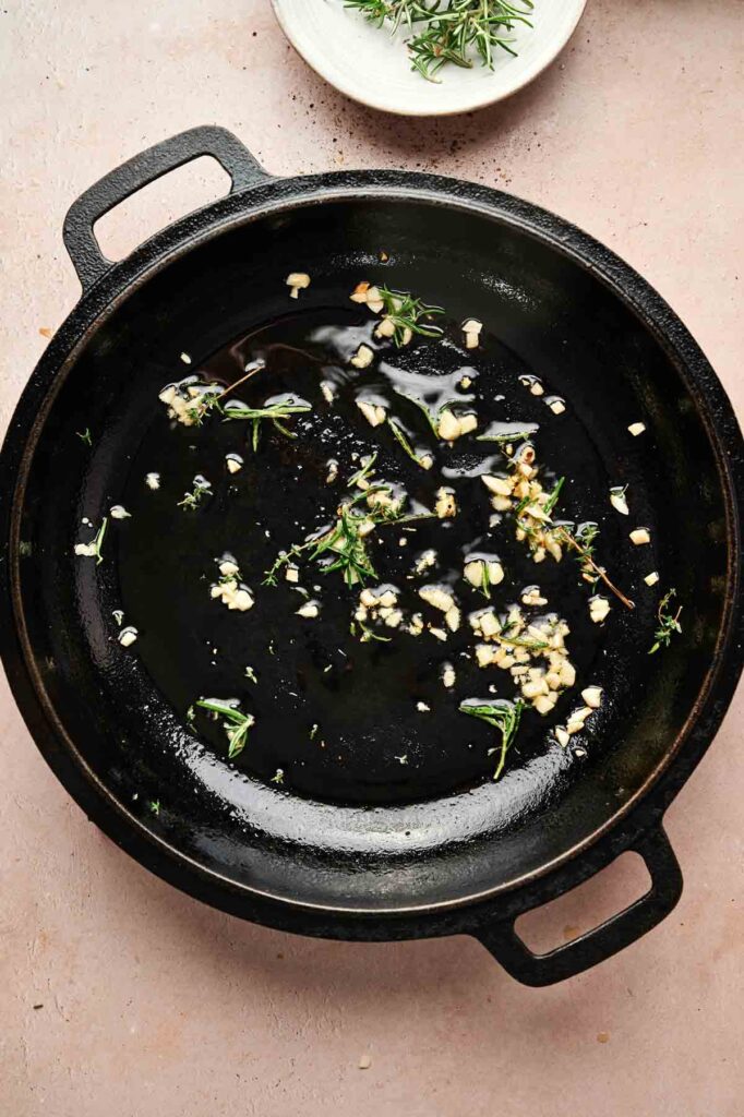 A black cast iron skillet with chopped garlic and fresh herbs sautéing in oil, perfect for starting a reverse sear steak, sits on a light countertop with a small bowl of herbs nearby.
