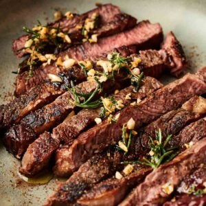Reverse Sear Steak sliced and topped with chopped garlic and fresh herbs, served elegantly on a grey plate.