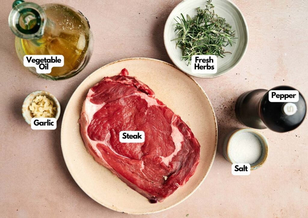 A raw steak prepared for a Reverse Sear Steak is on a plate, surrounded by vegetable oil, chopped garlic, fresh herbs, salt, and a pepper grinder on a light surface.