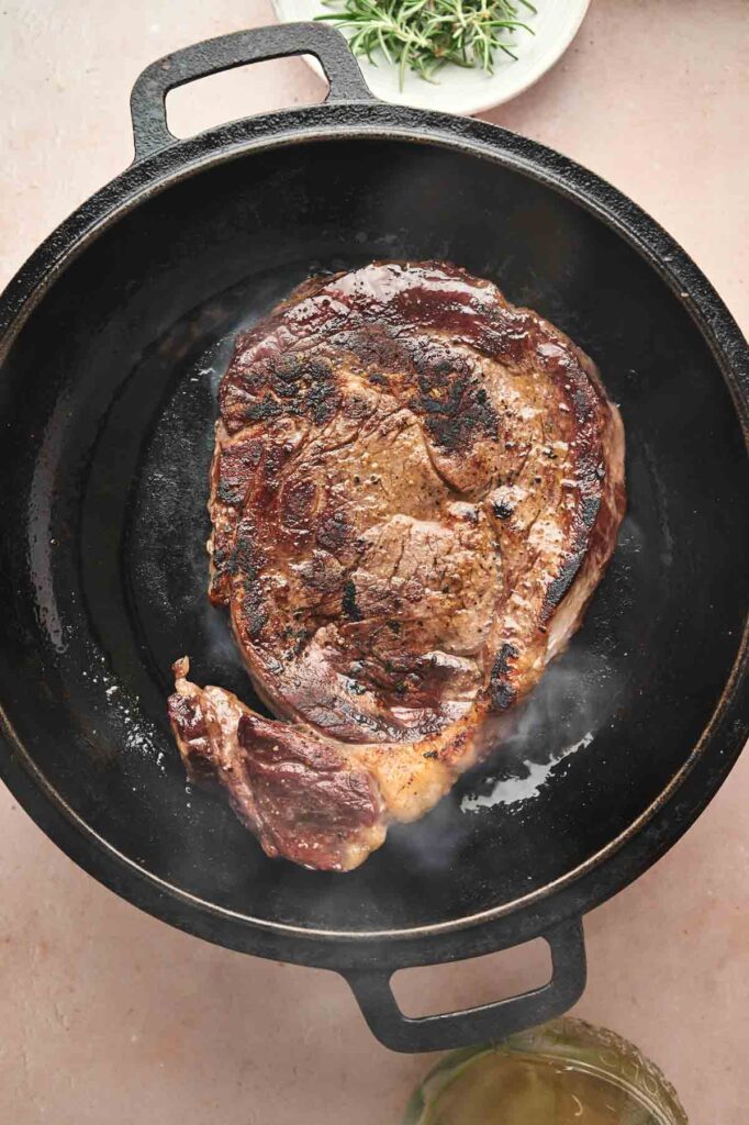 A Reverse Sear Steak is cooking in a black cast iron skillet, with steam rising and a sprig of rosemary visible on a plate in the background.
