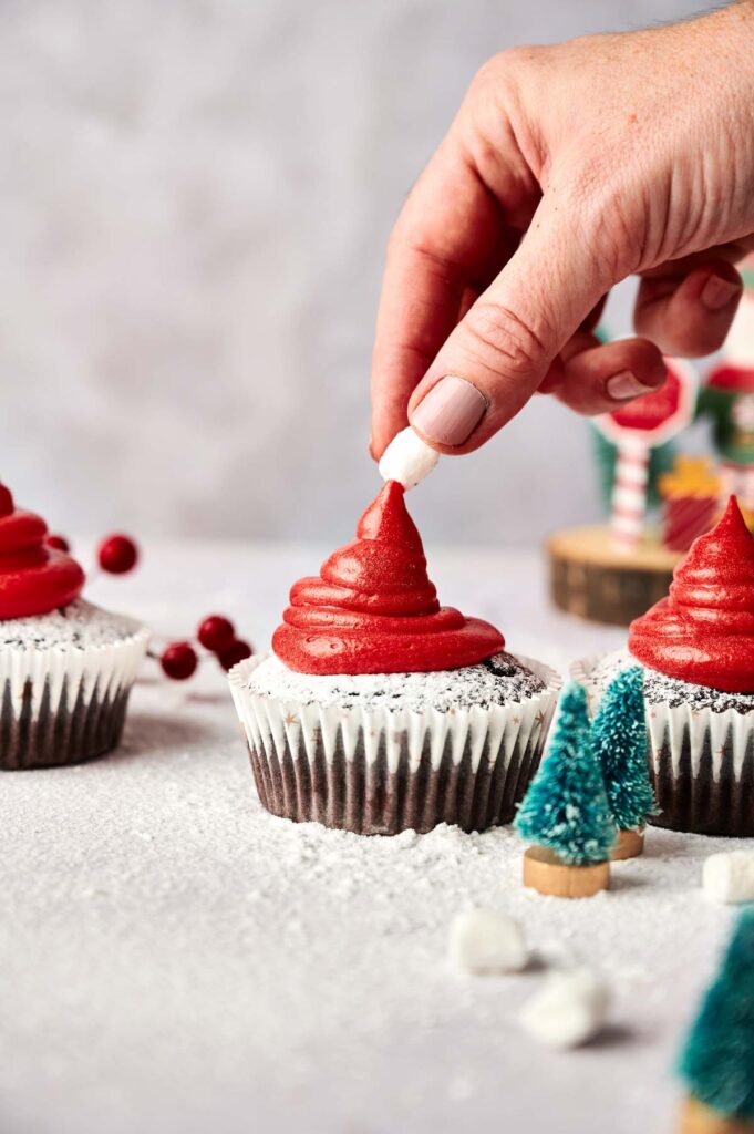 A hand places a mini marshmallow on top of a Santa Cupcake with red frosting, while other cupcakes and small Christmas tree decorations are nearby.