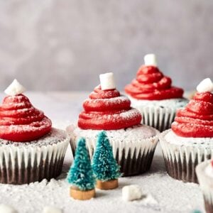 Four Santa Cupcakes decorated with red frosting and marshmallows to resemble Santa hats, surrounded by small artificial pine trees and a dusting of powdered sugar.