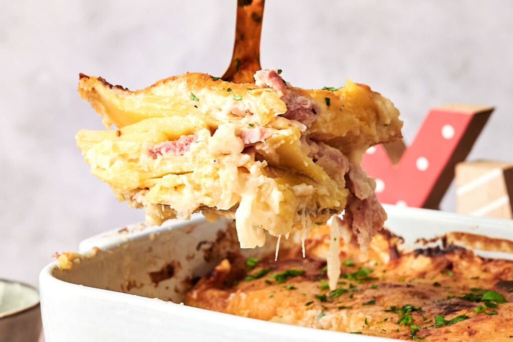A close-up of a serving of baked pasta with ham and cheese being lifted from a casserole dish, its creamy, golden-brown top reminiscent of classic Scalloped Potatoes and Ham.