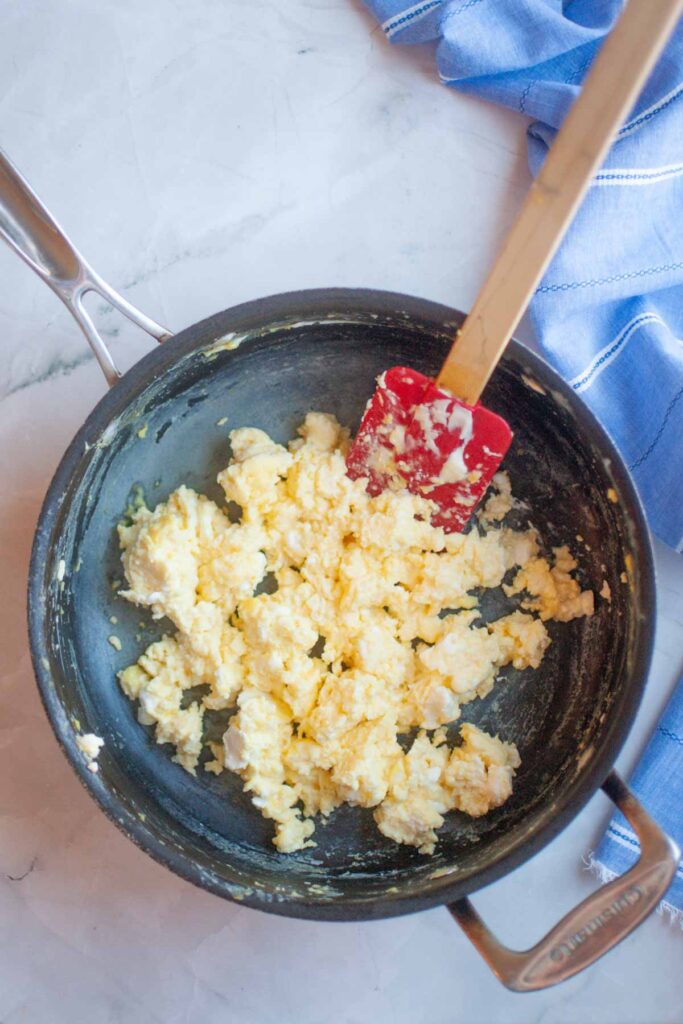 Scrambled Eggs with Cream Cheese are being cooked in a nonstick pan with a red spatula on a light countertop next to a blue cloth.
