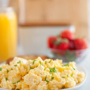 Plate of Scrambled Eggs with Cream Cheese garnished with chopped chives, with a bowl of strawberries and a glass of orange juice in the background.